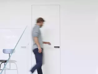 Das Bild zeigt einen Mann, der zu einer Tür läuft und sie öffnet. Auf der Tür ist der Griffwerk Türgriff R8 One in graphitschwarz zu sehen.