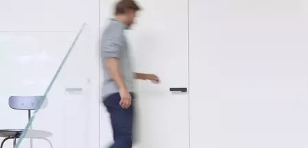 L'image montre un homme qui se dirige vers une porte et l'ouvre. Sur la porte, on peut voir la poignée de porte R8 ONE de Griffwerk en Noir graphite.