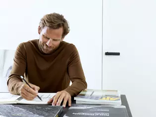 Un hombre se sienta concentrado en la mesa de su despacho y dibuja formas de asas. Disfruta dibujando, trabaja concentrado y puede disfrutar del tiempo para sí mismo.