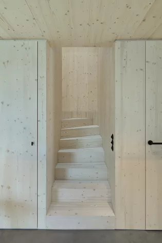 Die Abbildung zeigt eine Treppe aus hellem Holz, welche sich zwischen zwei Wänden, aus dem selben Holz befindet. An der Seite ist eine Tür mit dem schwarzen Türgriff LUCIA zu sehen. 