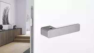 The picture shows the R8 One door handle by Griffwerk on a white door with a hallway in the background.