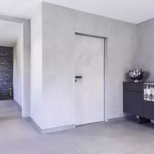 The illustration shows the plain entrance area in concrete look. From there, a Vitadoor Modulwerk 1.0. door with black anodized frame and the Frame door handle leads into the interior of the house. 