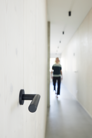 Lucia smart2lock in graphitschwarz Seitenansicht Die Abbildung zeigt den Griffwerk Türgriff Lucia smart2lock in graphitschwarz von der Seite. Im Hintergrund kommt eine Frau den Flur entlang gelaufen.