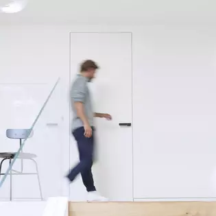 L'homme court vers la porte L'image montre un homme qui se dirige vers une porte et l'ouvre. Sur la porte, on peut voir la poignée de porte R8 ONE de Griffwerk en Noir graphite.