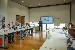 The illustration shows a room where children sit at tables and listen intently to a woman talk about Plant for the Planet.