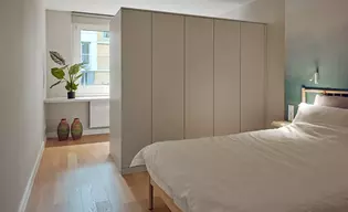 Die Abbildung zeigt ein Schlafzimmer. Im Vordergrund ist ein Bett, mit weißer Bettwäsche zu sehen, weiter hinten befindet sich ein großer, weißer Kleiderschrank, welcher den Raum von einem anderen Raum abtrennt.