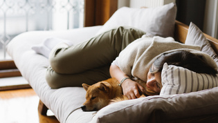 Die Abbildung zeigt eine Frau mit einem Hund zur Entspannung auf einer Couch liegen.