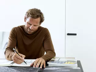 Mann sitzt konzentriert an seinem Bürotisch und zeichnet Griffformen. Er hat Freude am Zeichnen, arbeitet konzentriert und kann die Zeit für sich selbst genießen.