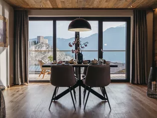 L'illustration montre la table de salle à manger dressée avec une baie vitrée. A travers celle-ci, on peut voir un château et les montagnes. 