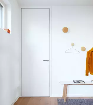  The picture shows a white door with the Griffwerk door handle Lucia smart2lock. Next to the door hangs a jacket and a hanger. Below is a bench on which lies a cloth.