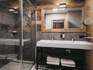 La foto muestra el cuarto de baño de un piso de tres habitaciones, con baldosas de mármol oscuro, acabados de madera y una ducha con paredes de cristal. 