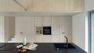 Die Abbildung zeigt im Vordergrund eine schwarze Küche. Im Hintergrund ist eine Holztreppe, Schränke und zwei Holztüren mit schwarzen Türgriffen ohne Schlüssellochrosette zu sehen.