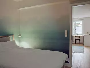 Die Abbildung zeigt ein Schlafzimmer mit einem Holzbett, mit weißer Bettwäsche und einer blau-verlaufenden Zimmerwand. Im Hintergrund ist ein lichter Durchgang zu einer Küche und Esszimmer zu sehen.
