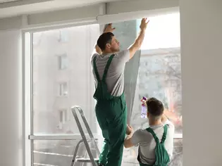 Das Bild zeigt zwei Handwerker, die ein Fenster mit Folie abdunkeln.