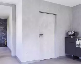 The illustration shows the plain entrance area in concrete look. From there, a Vitadoor Modulwerk 1.0. door with black anodized frame and the Frame door handle leads into the interior of the house. 