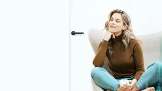 L'illustration montre une femme assise dans un fauteuil avec sa tasse de café. La porte derrière elle est fermée par la poignée de porte Lucia smart2lock en Noir graphite.