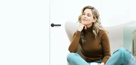 Die Abbildung zeigt ein Frau, die mit ihrer Kaffeetasse in einem Sessel sitzt. Die Tür hinter Ihr ist mit dem Griffwerk Türgriff Lucia smart2lock in graphitschwarz verschlossen.