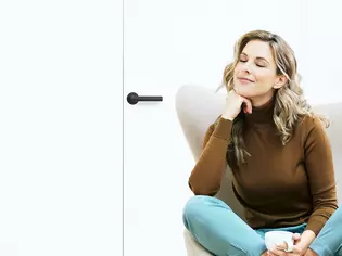 The picture shows a woman sitting in an armchair with her coffee cup. The door behind her is closed with the Griffwerk door handle Lucia smart2lock in graphite black.