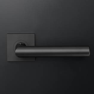 The picture shows the door handle Lucia in graphite black with a square rosette, mounted on a black wooden door.