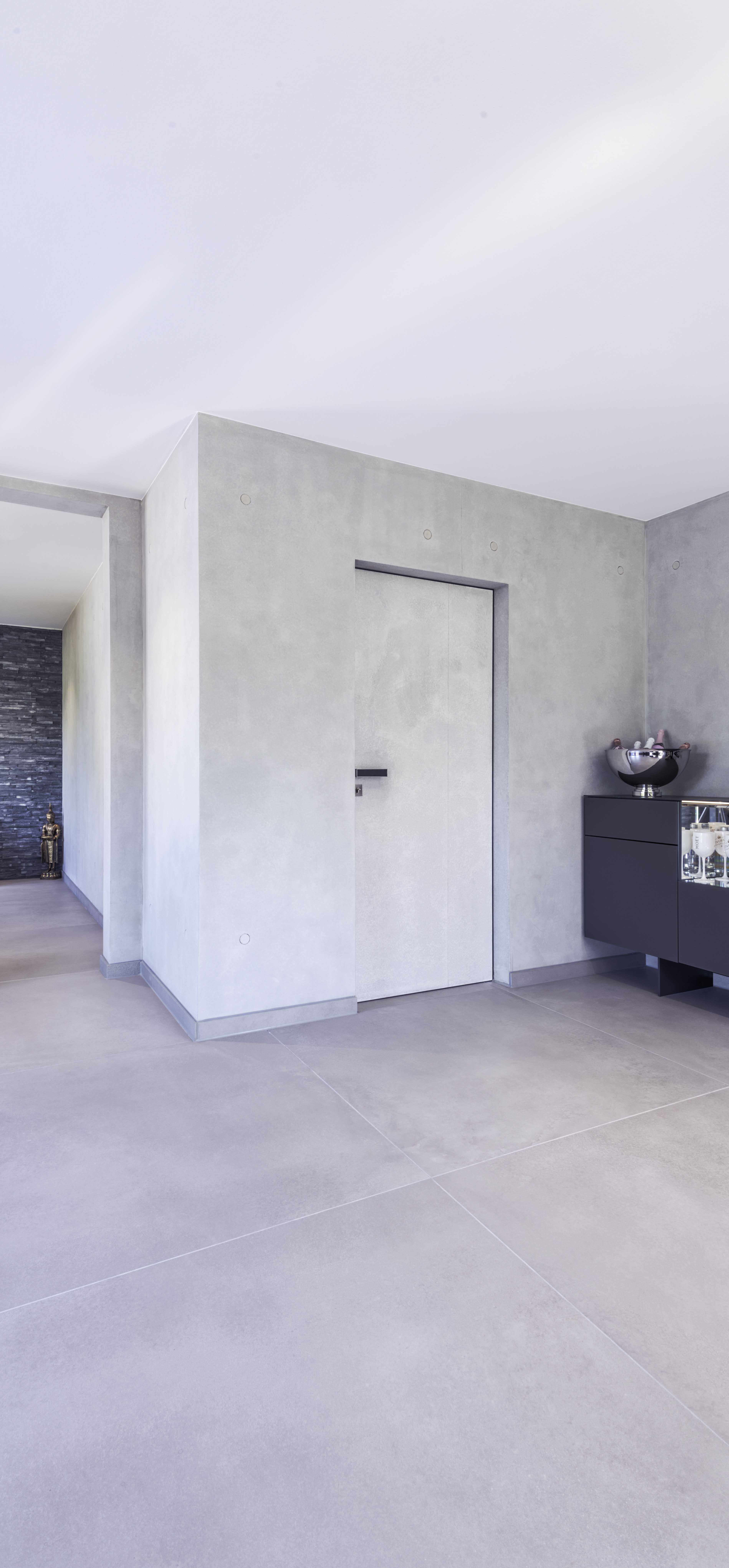 The illustration shows the plain entrance area in concrete look. From there, a Vitadoor Modulwerk 1.0. door with black anodized frame and the Frame door handle leads into the interior of the house. 